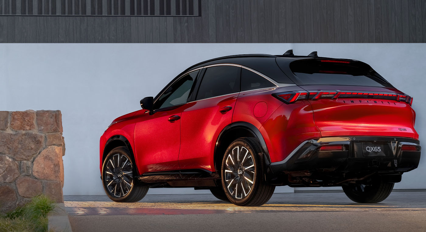 Red Infiniti QX65 SUV with a modern design is parked by a stone wall. The sun casts a warm glow on its glossy finish, highlighting the curves and detailing.